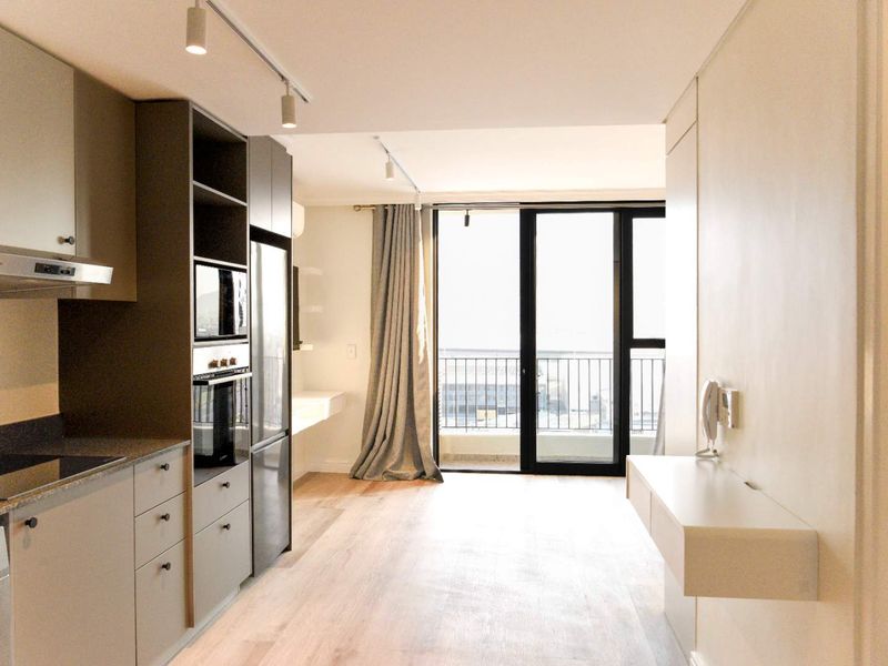 Modern open-plan apartment featuring a sleek kitchen with integrated appliances, light wood flooring, and direct access to a private balcony through sliding glass doors.