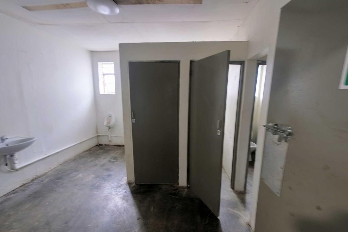 Industrial-style communal bathroom facility featuring a wall-mounted basin, urinal, and multiple partitioned toilet cubicles with basic finishes.