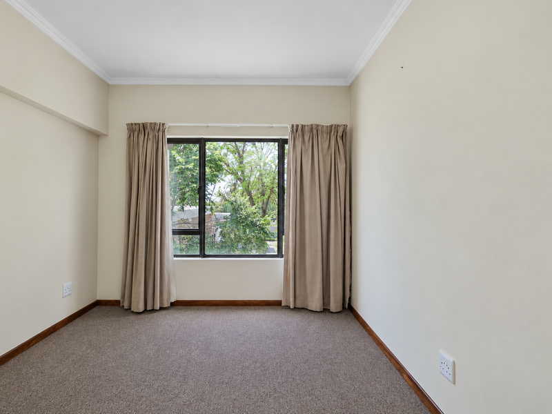 A bright, empty bedroom featuring neutral walls, carpeted flooring, and a large window with curtains overlooking greenery.