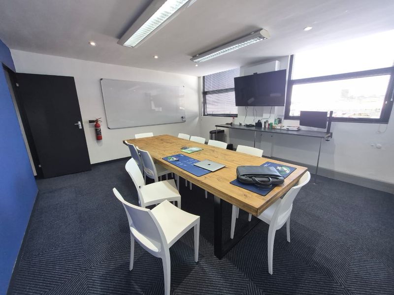 A functional office meeting room featuring a large central table, wall-mounted whiteboard, television, and integrated desk space with modern carpet flooring.