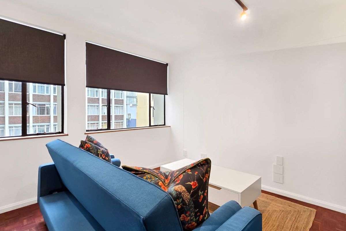 A bright living room featuring classic parquet flooring, track lighting, and large windows with fitted roller blinds.