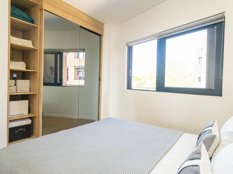 A bright bedroom featuring a built-in wardrobe with mirrored sliding doors, open shelving, and a large window with a roller blind.