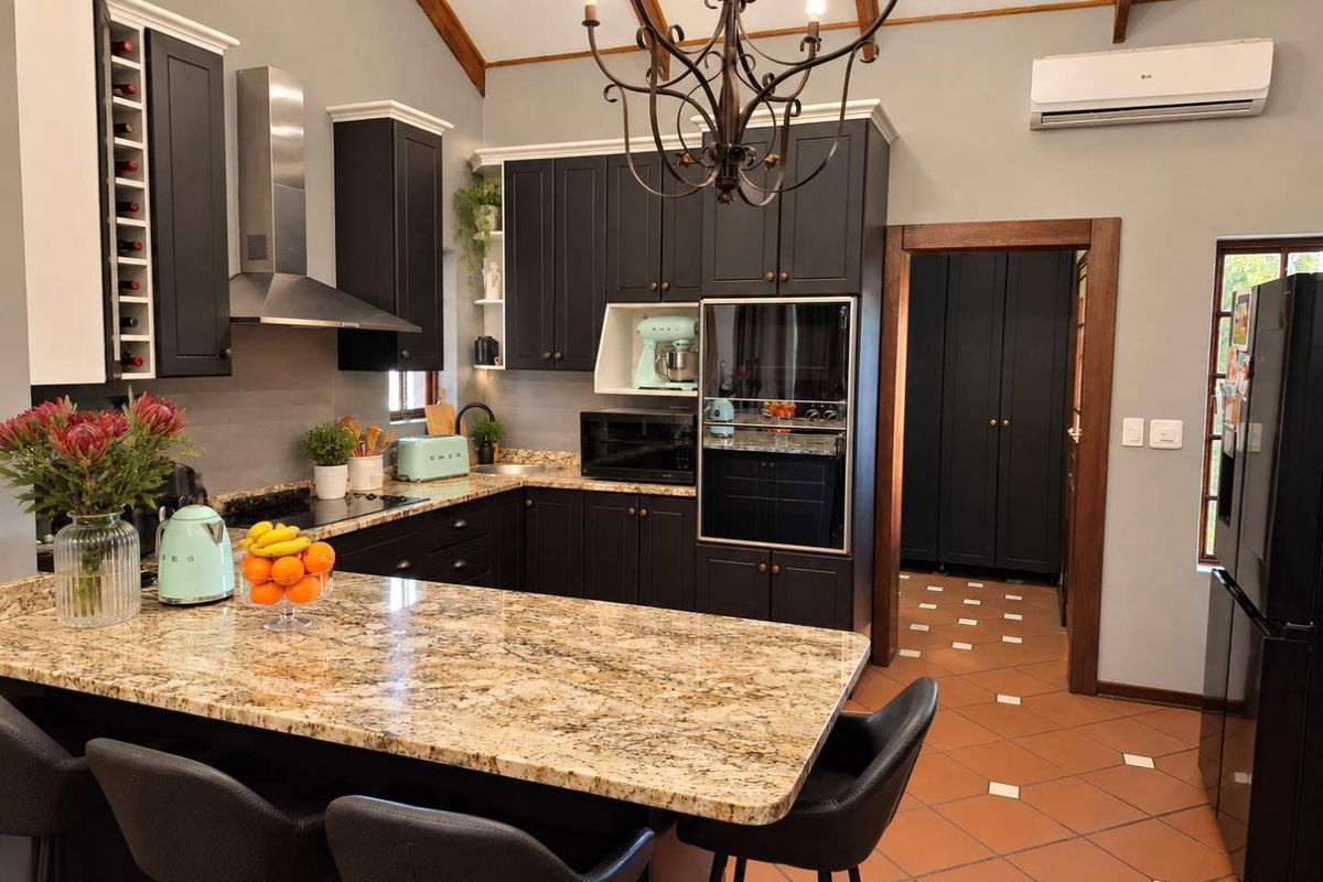Modern kitchen featuring dark cabinetry, granite countertops, vaulted ceilings with exposed beams, and integrated appliances, leading into a tiled utility or pantry area.