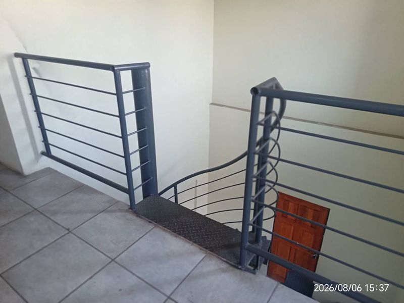 An interior view of a staircase landing featuring metal railings and tiled flooring, leading down to a lower level with a wooden door.