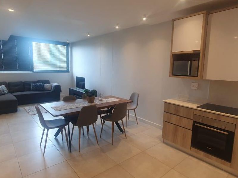 Modern open-plan living area featuring a contemporary kitchen with built-in appliances, tiled flooring, and recessed lighting, seamlessly flowing into the dining and lounge space.