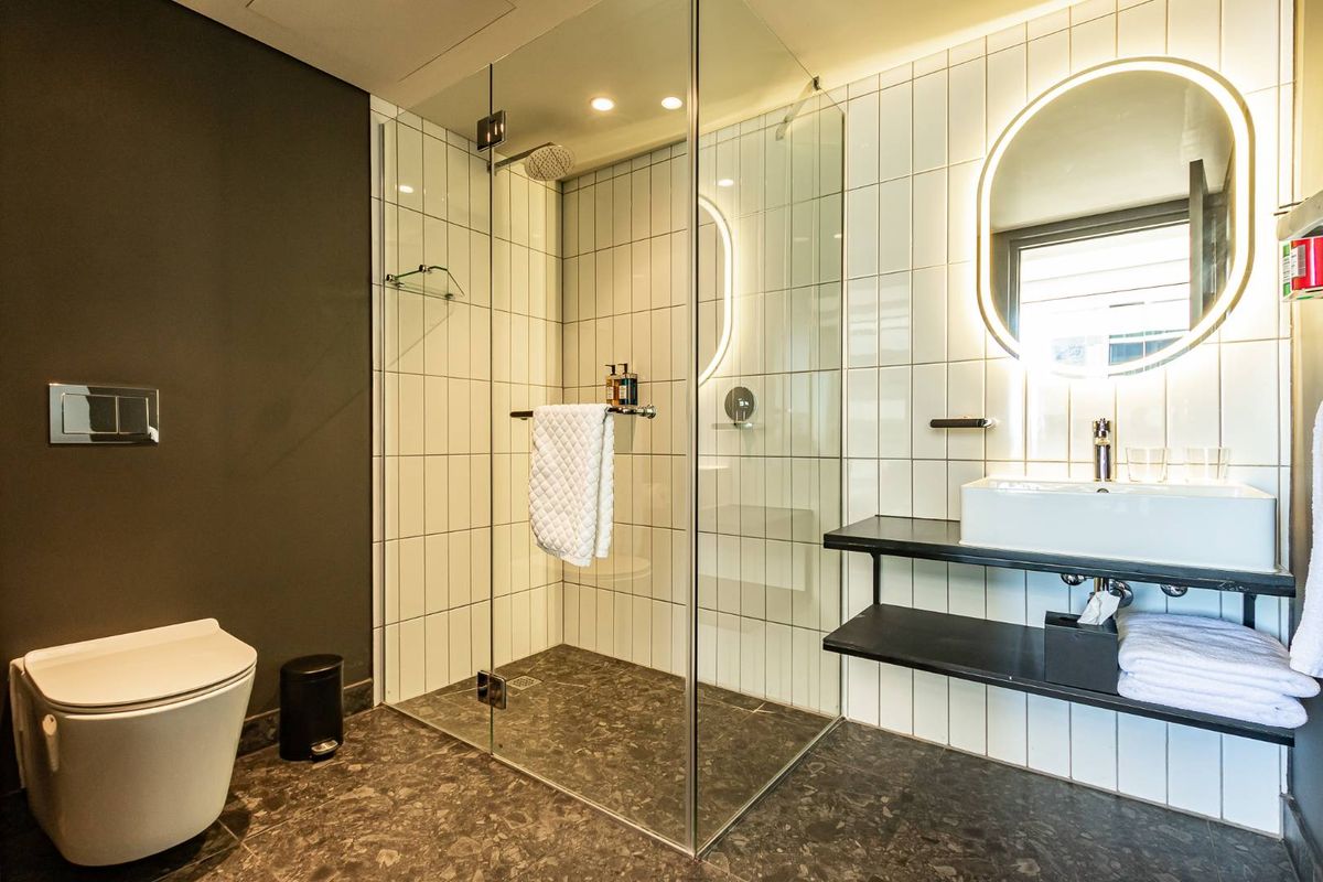 Modern bathroom featuring a glass-enclosed walk-in shower, wall-mounted toilet, vessel sink on a floating shelf, and illuminated circular mirror.