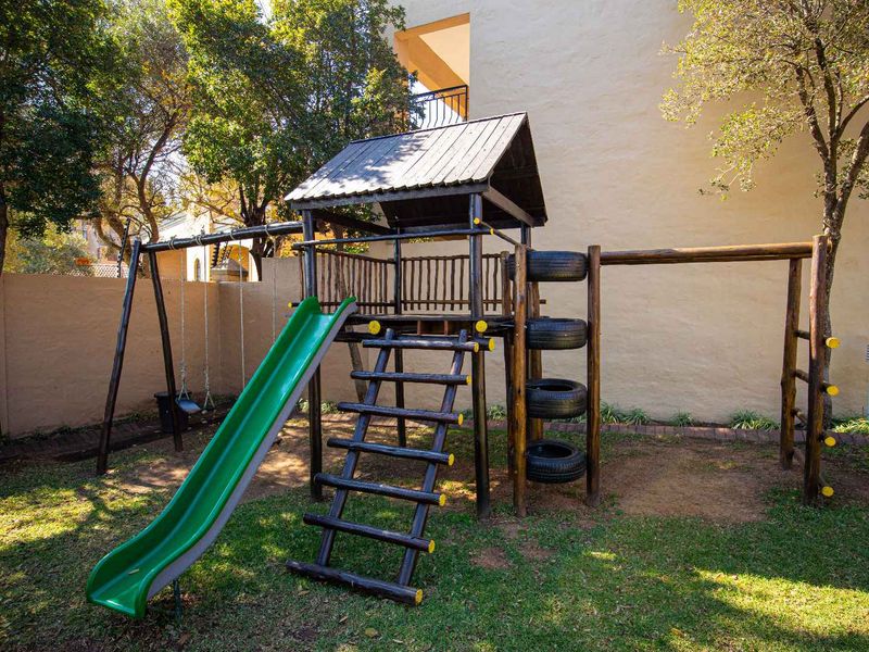 A communal garden area featuring a wooden jungle gym with a slide, swing set, and climbing structure, set against a residential building wall.