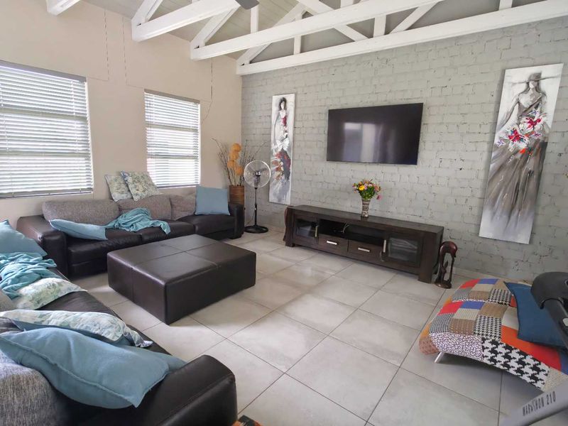 Spacious living room featuring a painted brick accent wall, exposed timber roof trusses, and neutral tiled flooring with large windows and venetian blinds.