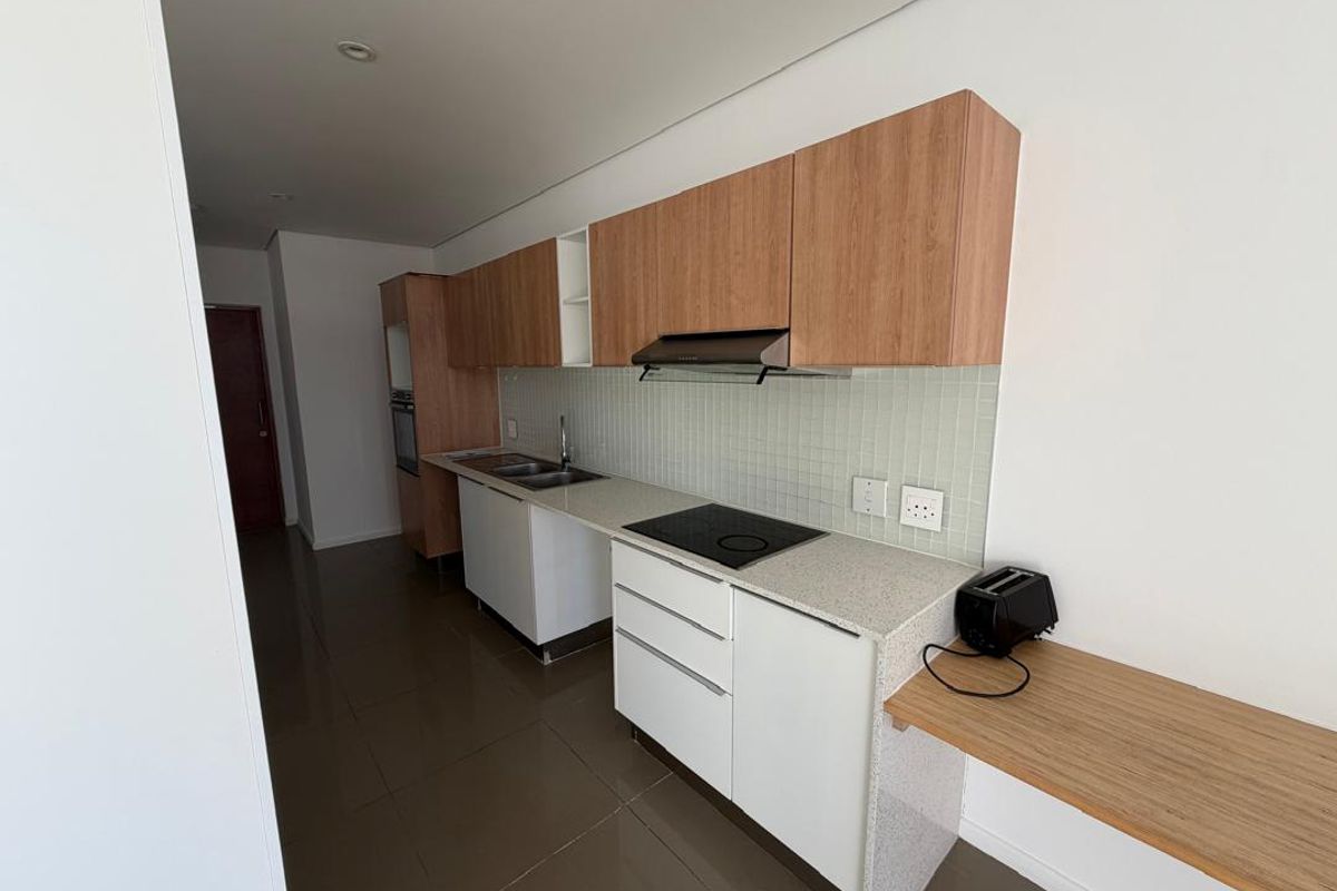 Modern galley-style kitchen featuring light wood-tone cabinetry, stone countertops, integrated electric hob, extractor fan, and sleek tiled backsplash.