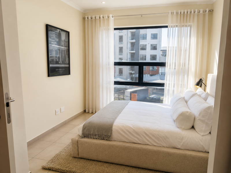 A well-lit bedroom featuring tiled flooring, neutral wall tones, and a large window with sheer curtains, offering a clean and comfortable living space.