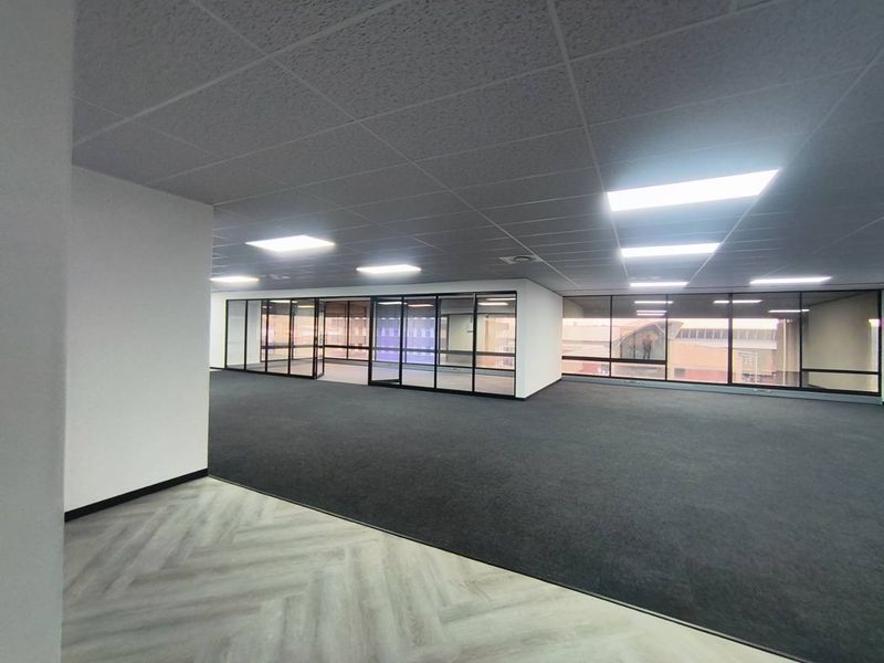 A spacious, modern open-plan office area featuring glass partitioning, a suspended ceiling with recessed lighting, and a combination of carpet and vinyl flooring.