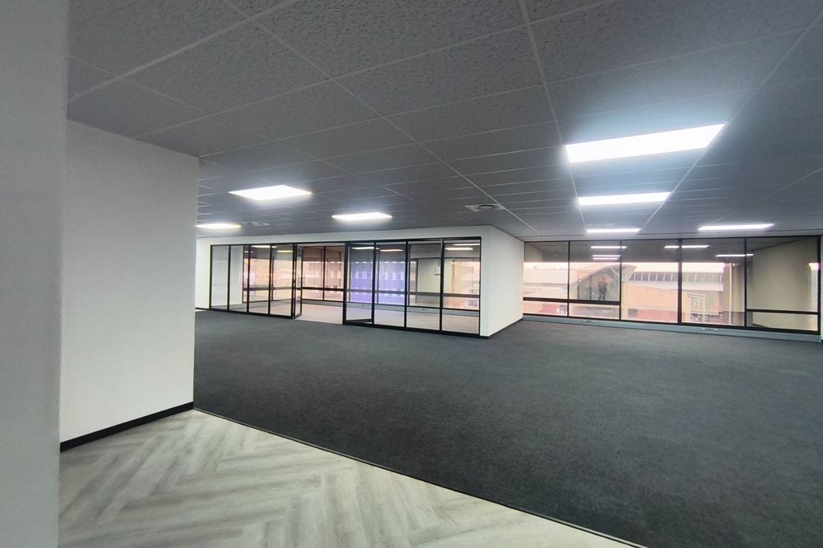 A spacious, modern open-plan office area featuring glass partitioning, a suspended ceiling with recessed lighting, and a combination of carpet and vinyl flooring.