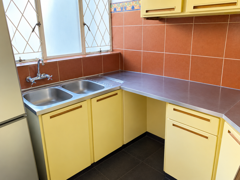 A functional kitchen area featuring a stainless steel double sink, metal countertops, and dated cabinetry with a tiled backsplash and leaded glass window.