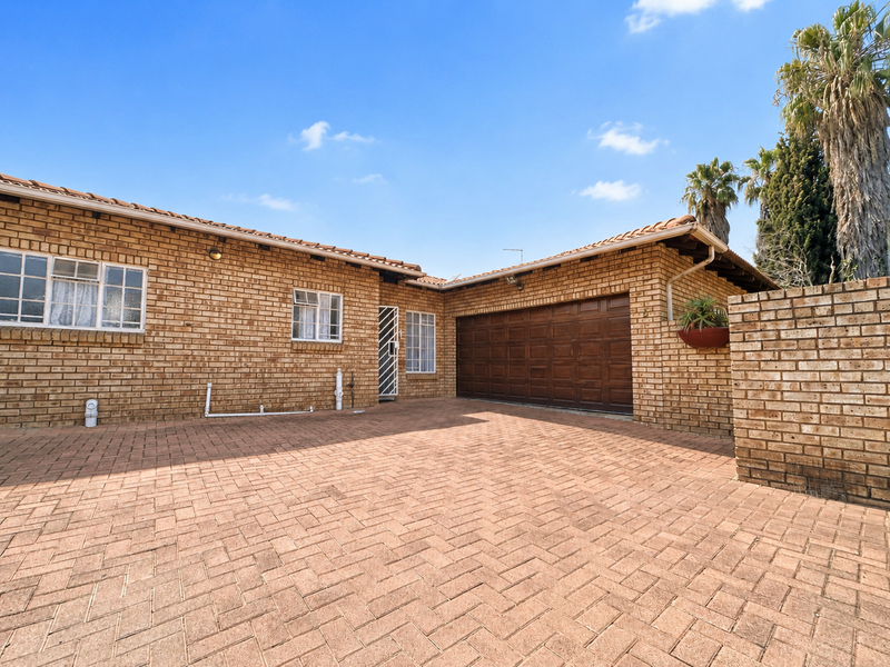 Front exterior view of a brick house featuring a paved driveway, double garage, and secure entrance gate under a clear blue sky.