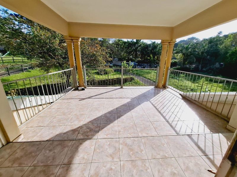 A spacious covered balcony featuring tiled flooring, decorative columns, and a metal balustrade, offering an open view of the surrounding garden area.