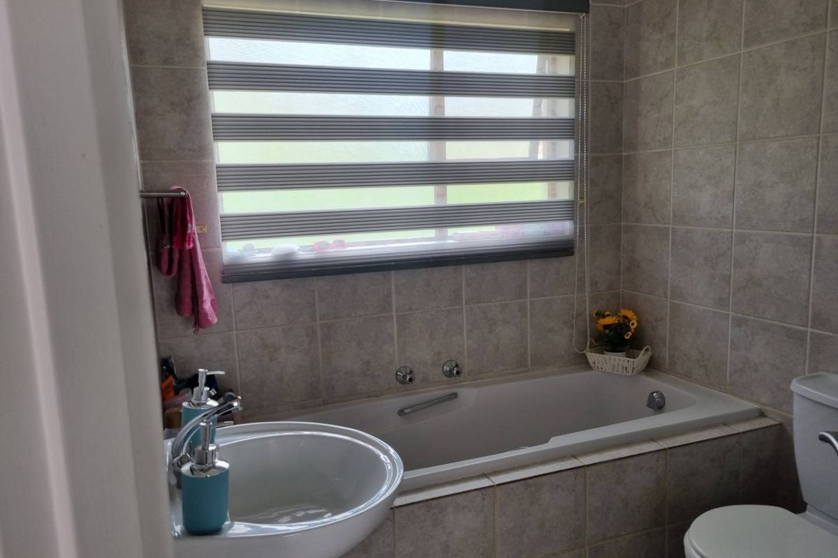 A functional bathroom featuring a built-in bathtub, pedestal sink, toilet, and neutral wall tiling with a window for natural light.