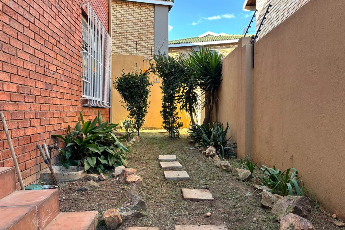 A narrow side garden area featuring a brick facade, paved stepping stones, and perimeter boundary walls with security fencing.