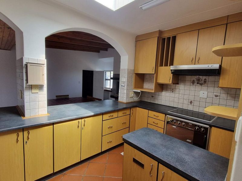 A functional kitchen featuring wooden cabinetry, laminate countertops, an integrated stove with extractor fan, and terracotta floor tiles, connected to the living area via archway.
