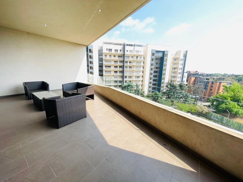 A spacious covered balcony featuring tiled flooring, recessed lighting, and a modern glass balustrade with views of the surrounding apartment complex.