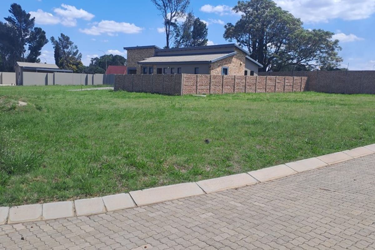A vacant residential plot featuring a paved driveway, established grass lawn, and brick boundary walling, offering a clear space for future development.