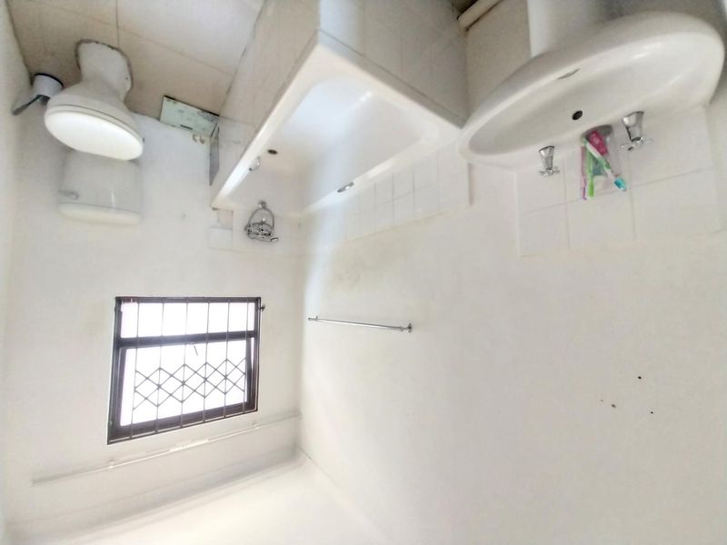 A dated bathroom featuring a bathtub, pedestal basin, and toilet, with security bars on the window and visible signs of wear on the walls.