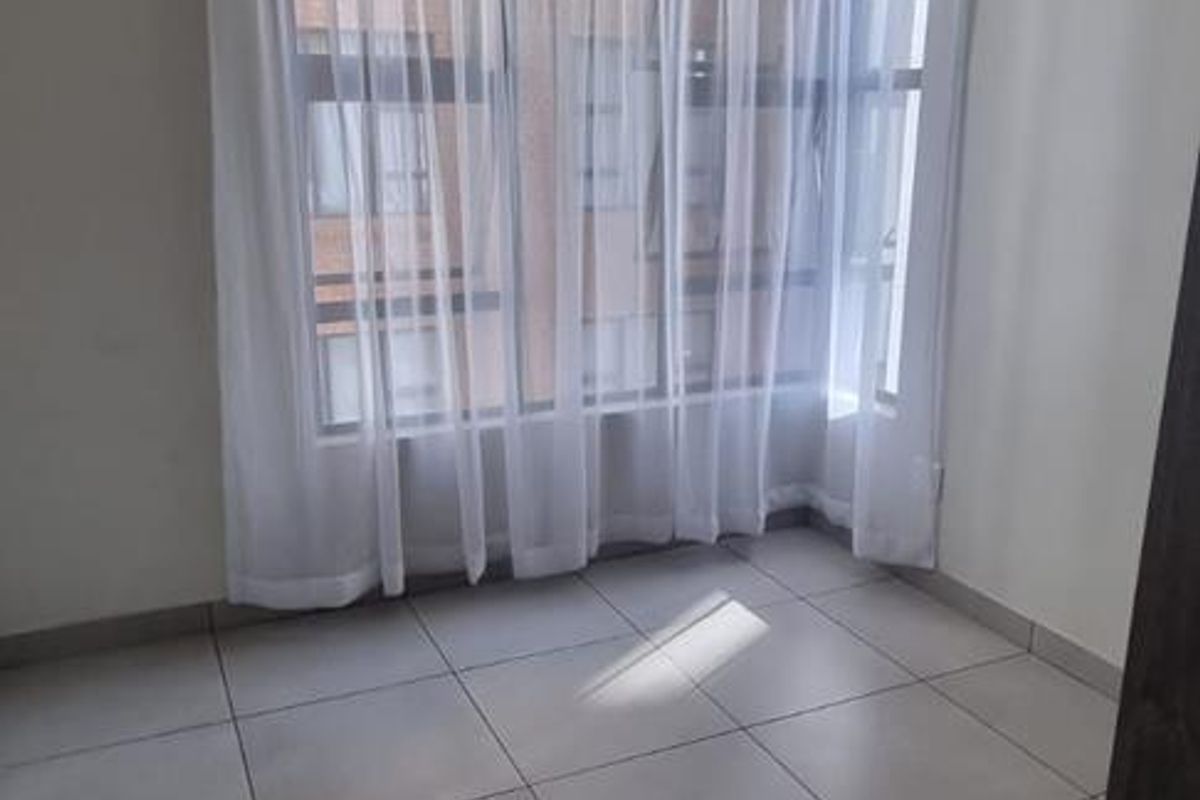 A bright, empty bedroom featuring large windows with sheer curtains and neutral floor tiling, offering a clean and versatile living space.
