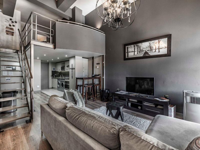 Modern open-plan apartment featuring a mezzanine level, metal staircase, and a compact kitchen with integrated appliances, finished with contemporary grey tones.