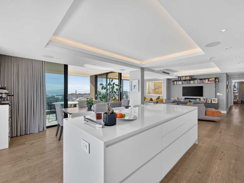 Modern open-plan living area featuring a sleek kitchen island, hardwood flooring, recessed ceiling lighting, and expansive sliding doors leading to a balcony with views.