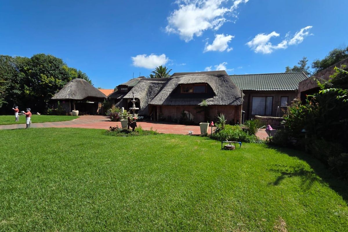 A spacious rear garden featuring a large lawn, paved patio area, and a traditional thatched-roof house structure.