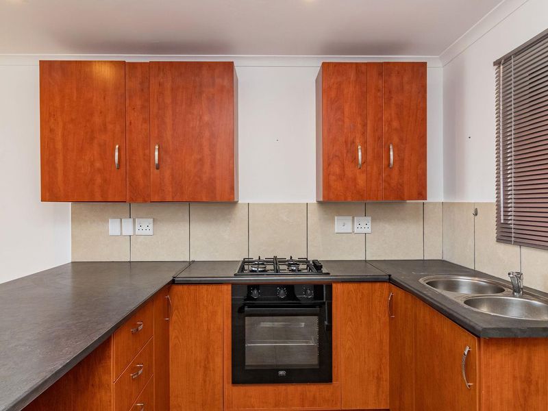 A functional kitchen featuring wood-finish cabinetry, dark laminate countertops, a built-in oven with gas hob, and a double bowl sink with tiled backsplash.