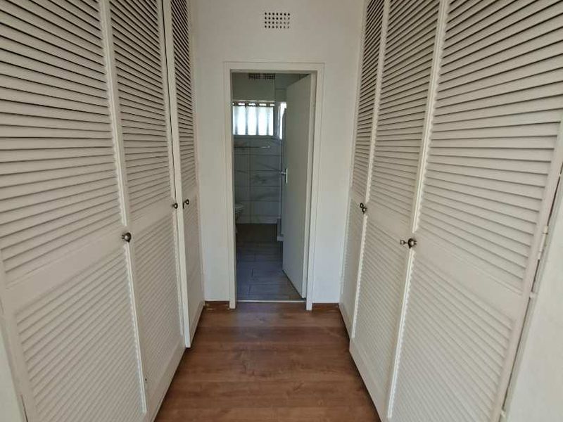 A hallway featuring extensive built-in louvered cupboards and laminate flooring, leading towards an adjacent bathroom area.