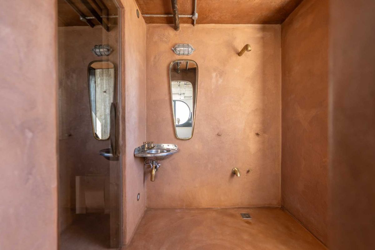 A unique, industrial-style bathroom featuring terracotta-toned plaster walls, brass fixtures, a wall-mounted basin, and a walk-in shower area.