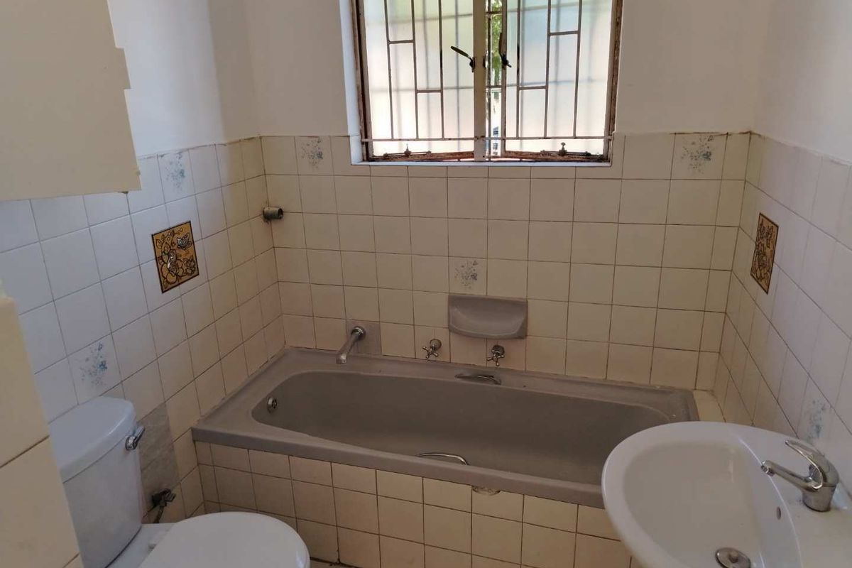 A dated bathroom featuring a built-in bathtub, pedestal basin, and toilet, with original wall tiling and security bars on the window.