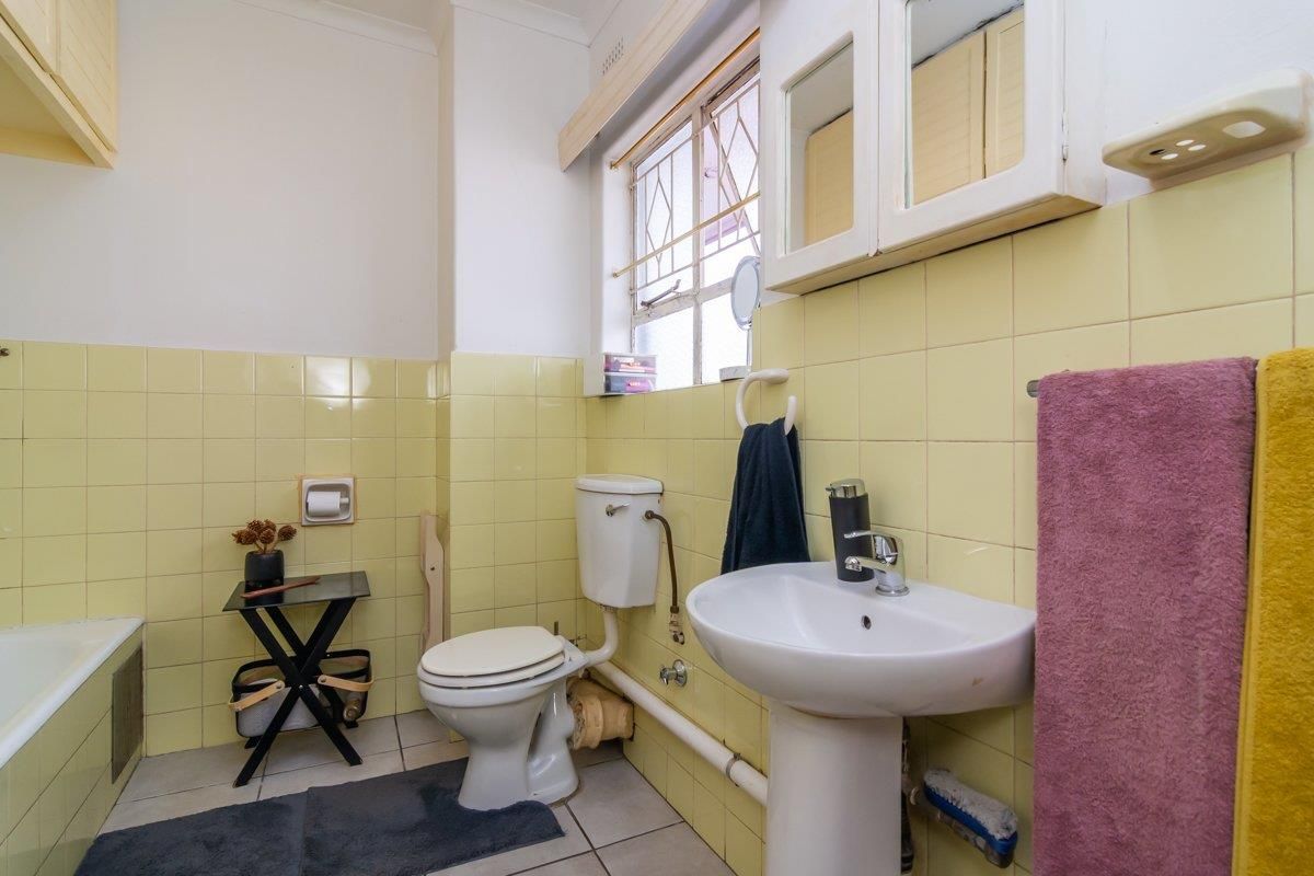 A dated bathroom featuring yellow wall tiles, a pedestal basin, a standard toilet, and a bathtub with a window providing natural light.