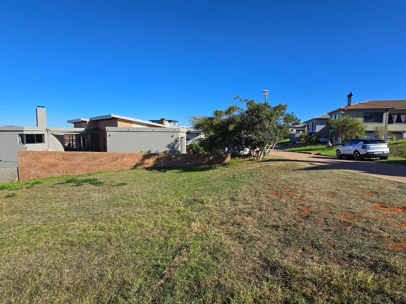 A view of a vacant residential plot featuring established grass, a brick boundary wall, and surrounding residential properties in a developed estate setting.