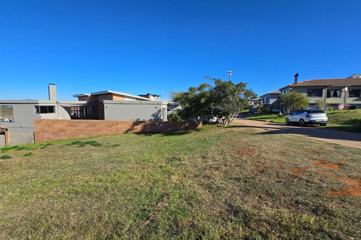 A view of a vacant residential plot featuring established grass, a brick boundary wall, and surrounding residential properties in a developed estate setting.