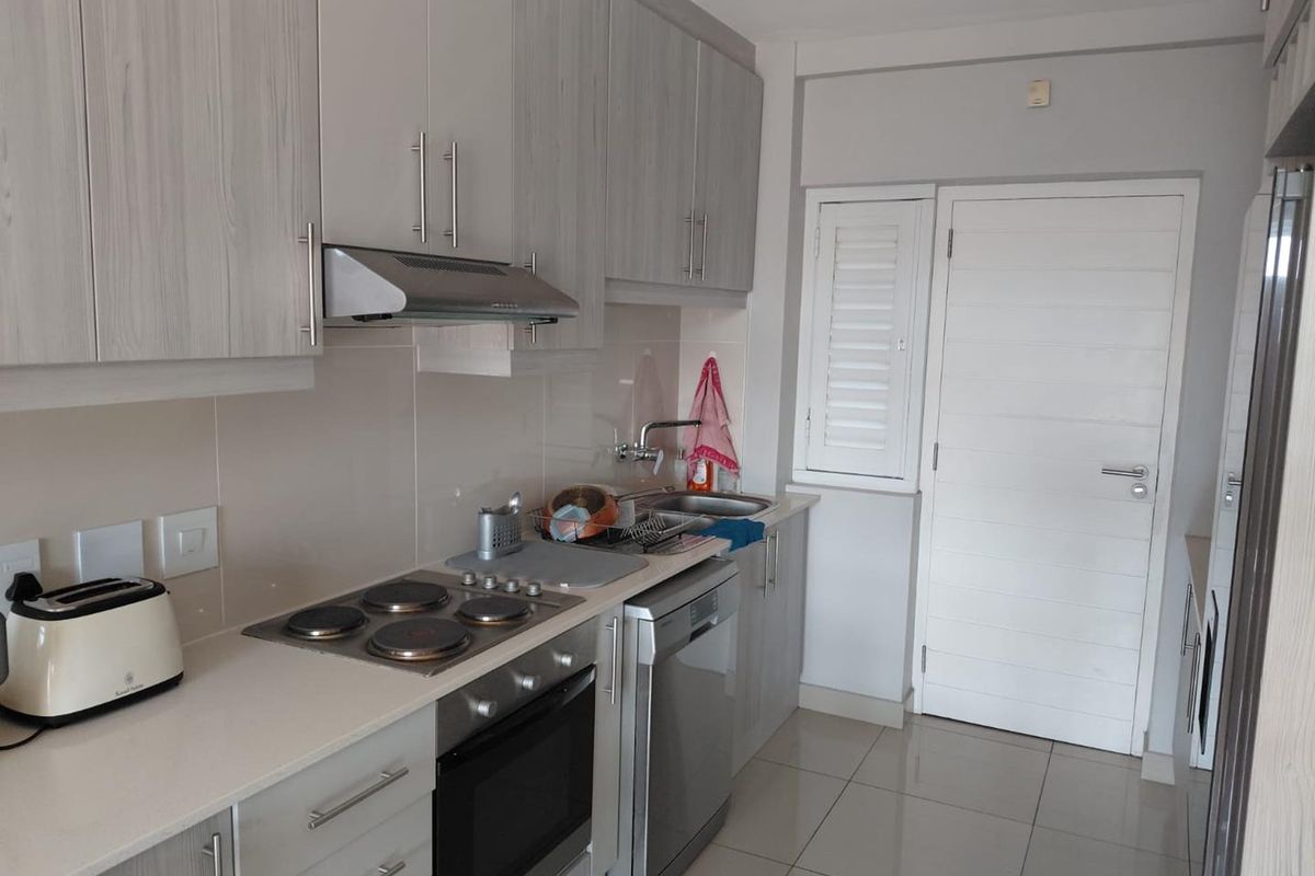 A functional kitchen featuring light wood-grain cabinetry, integrated electric hob and oven, stainless steel sink, and neutral tiled flooring.