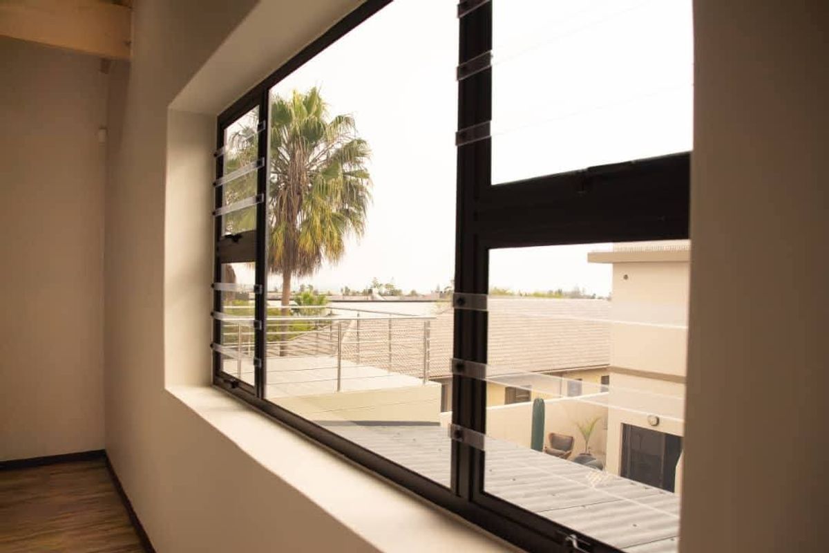 A bright, empty bedroom featuring modern aluminium window frames with security bars and laminate flooring, offering a view of the surrounding neighbourhood.