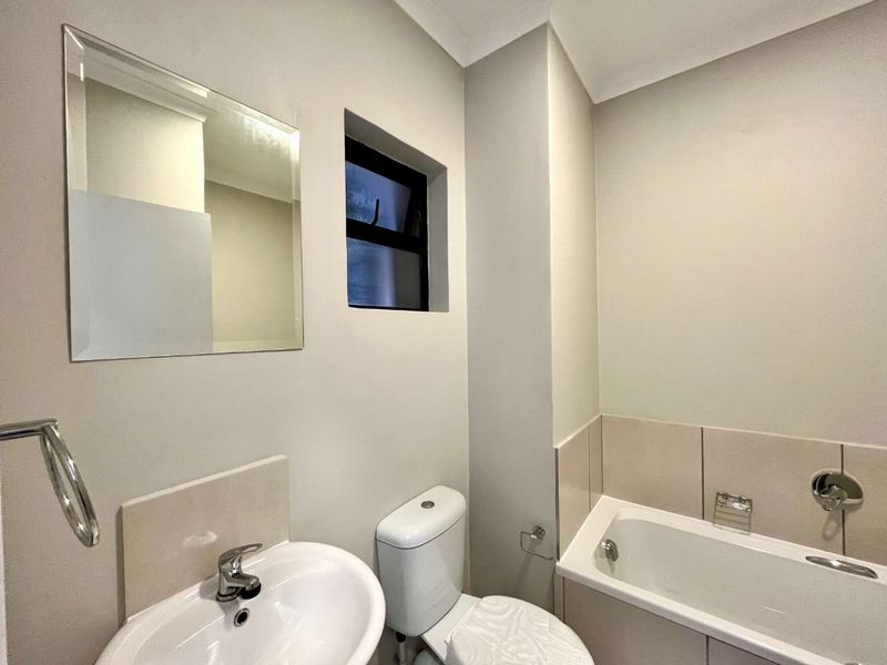 A clean, modern bathroom featuring a built-in bathtub, wall-mounted basin, toilet, and a wall mirror with neutral tiling.