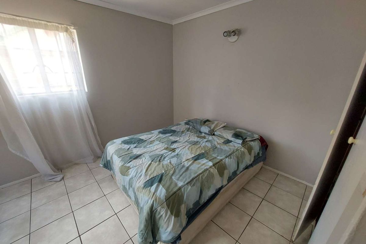 A simple bedroom featuring tiled flooring, a single window with curtaining, and a wall-mounted light fixture.