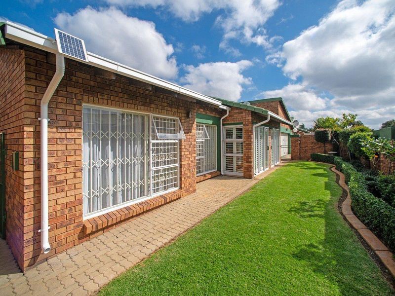 A well-maintained rear exterior view of a face brick cluster home featuring a paved walkway, manicured lawn, and secure window gates.