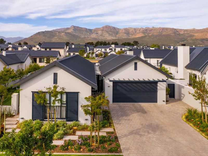 A modern residential property featuring a metal roof, paved driveway, double garage, and landscaped garden, set against a scenic mountain backdrop.