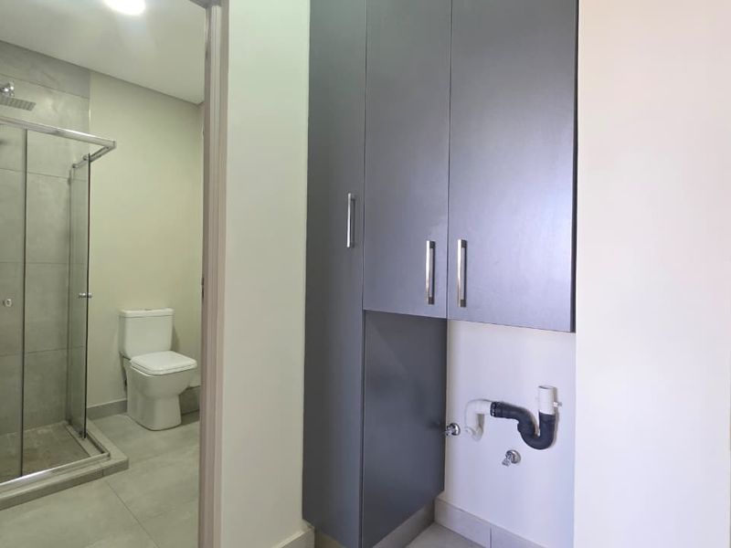 A functional laundry area featuring built-in cabinetry and plumbing connections, adjacent to a modern tiled bathroom with a shower and toilet.