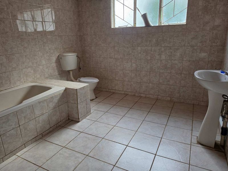 A dated bathroom featuring a built-in bathtub, pedestal basin, and toilet, finished with full-height ceramic wall tiling and floor tiles.