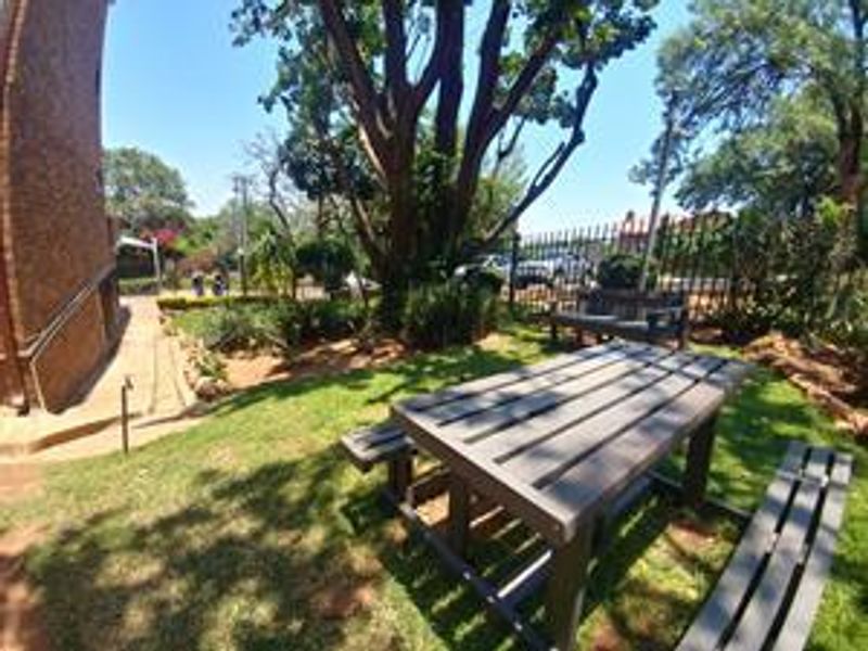 A well-maintained garden area featuring mature trees, a paved walkway along the brick building facade, and secure perimeter fencing.