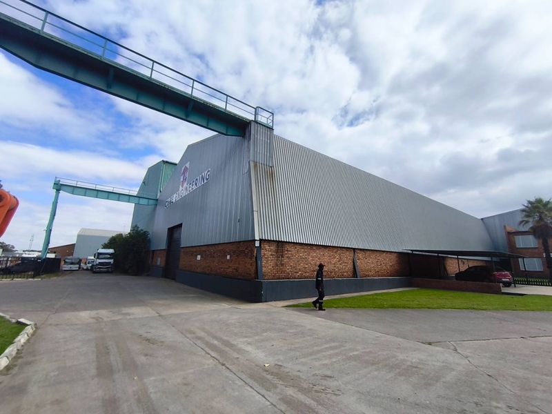 Industrial facility featuring corrugated metal cladding, brick masonry base, and an overhead gantry conveyor system with paved driveway access.