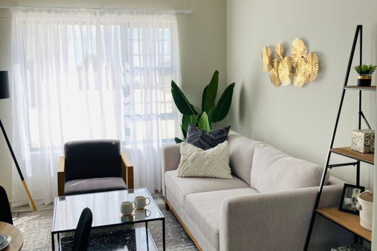 A well-lit living room featuring neutral wall tones, a large window with sheer curtains, and a functional layout suitable for comfortable seating.
