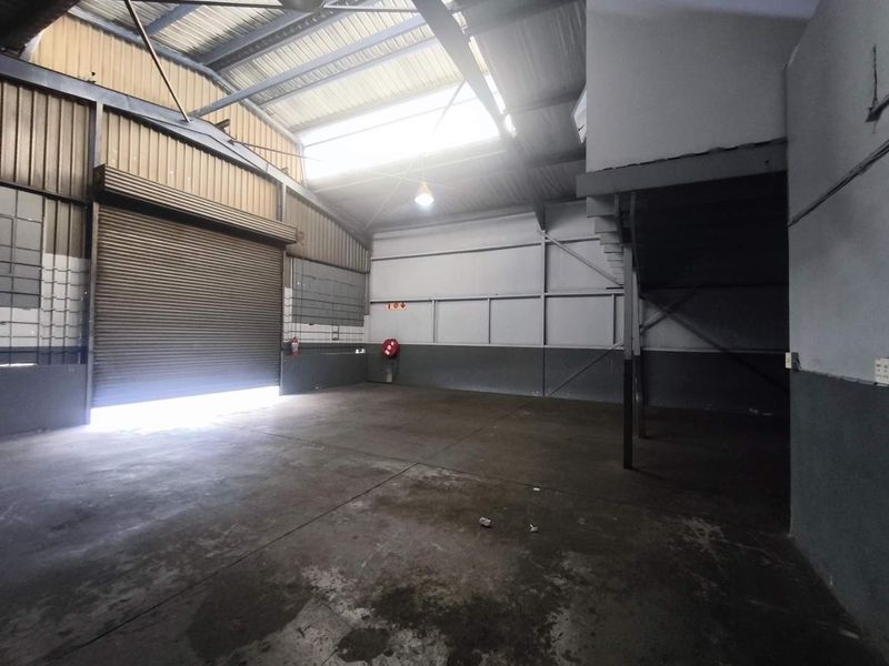 Spacious industrial utility area featuring a high-clearance roller shutter door, exposed steel roof trusses, a skylight, and a mezzanine level for additional storage or workspace.