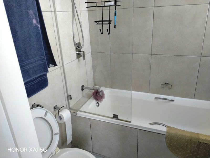A functional bathroom featuring a built-in bathtub with a glass shower screen, wall-mounted fixtures, and neutral ceramic wall tiling.