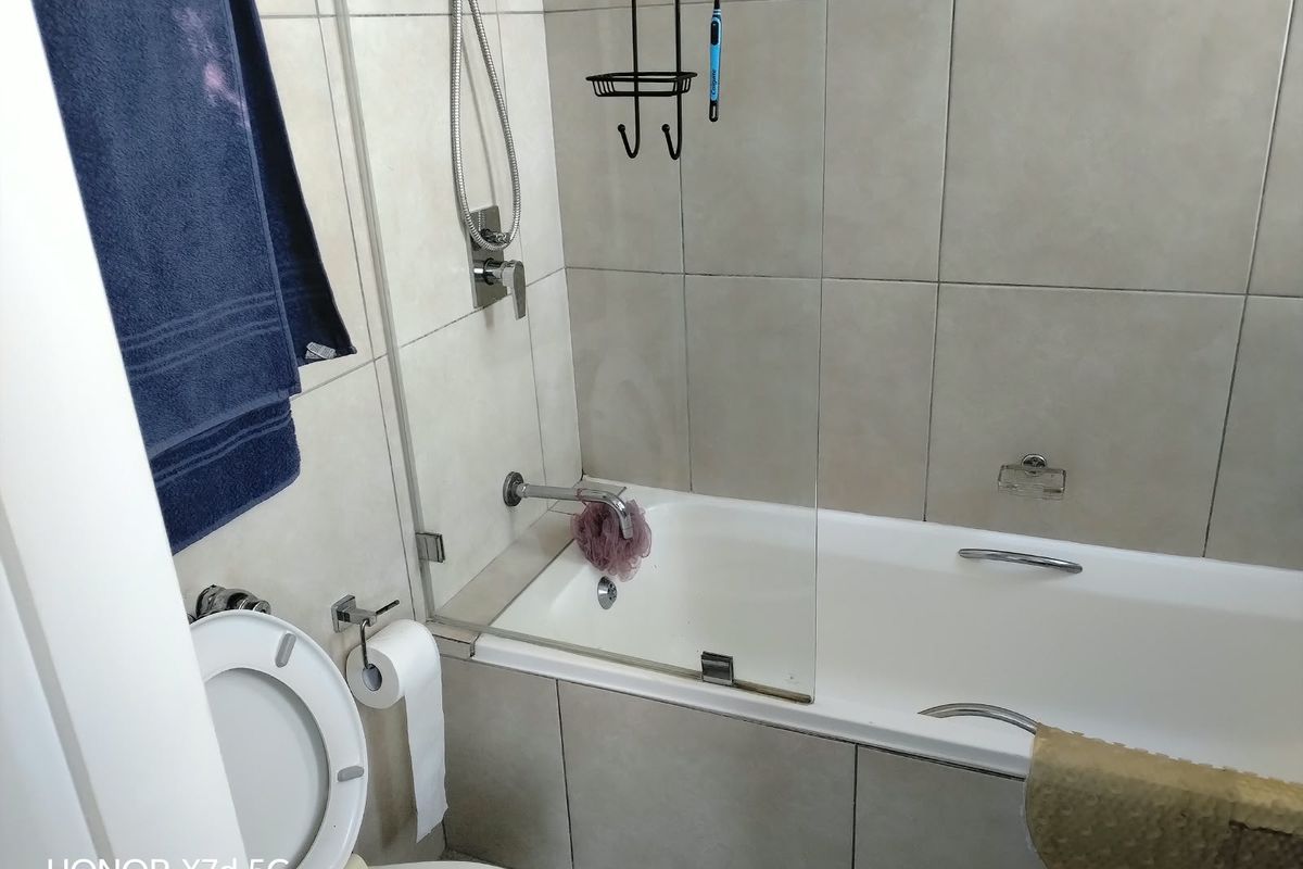 A functional bathroom featuring a built-in bathtub with a glass shower screen, wall-mounted fixtures, and neutral ceramic wall tiling.
